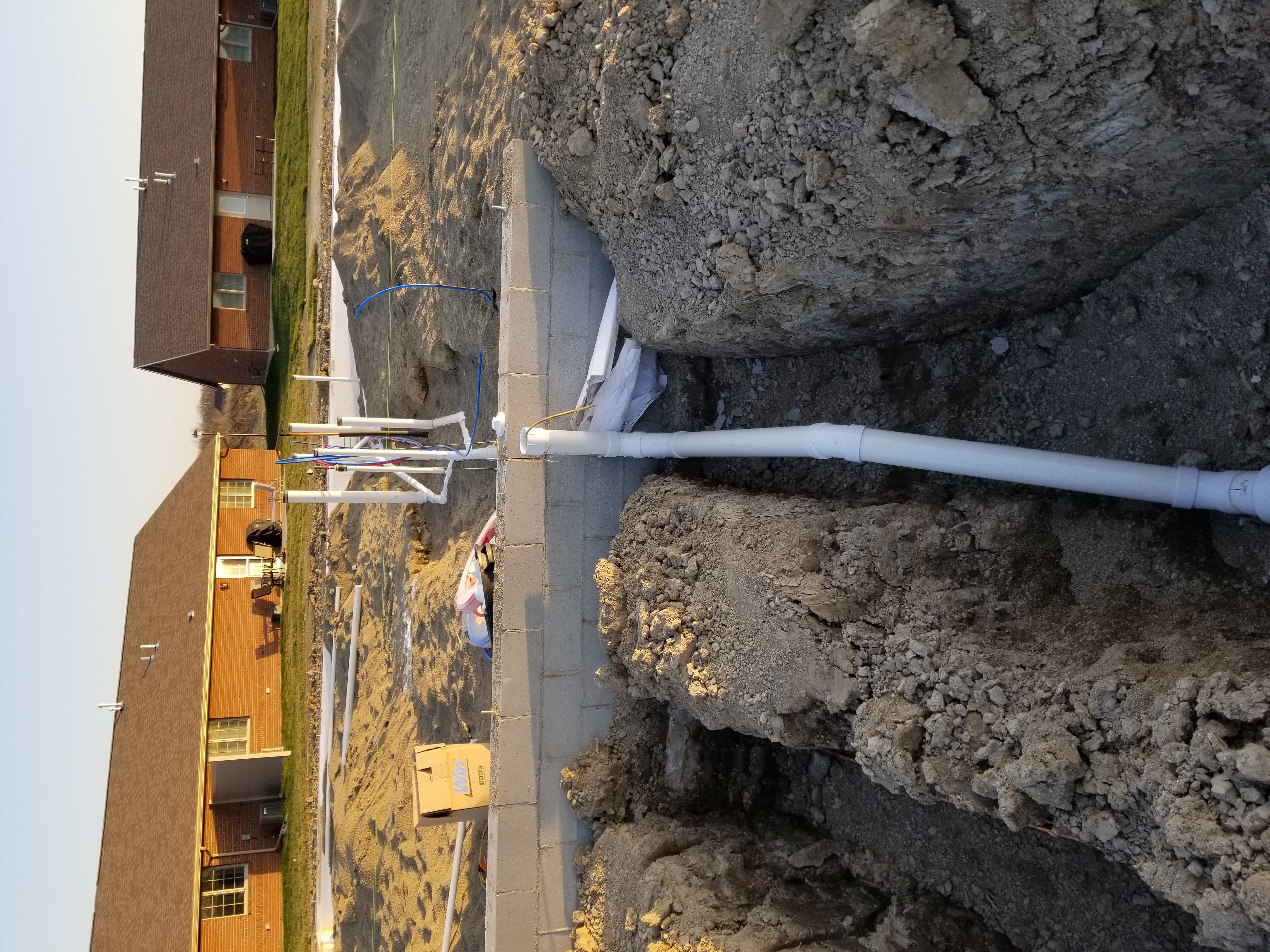 Plumbing pipes in trenches on a construction site, with dirt, gravel, and trees in the background